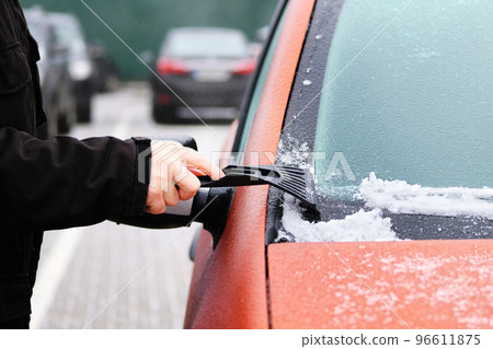 Man clears snow from icy windshield of car. Scraper in mans hand. Cleaning frozen window of orange automobile. 96611875