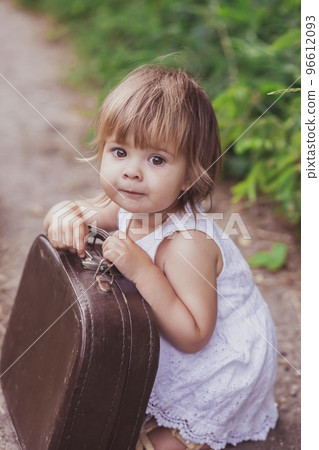 Charming baby in a white sundress tries to open an old suitcase 96612093