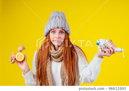 sick illness redhaired woman wearing knitted sweater,hat with scarf choosing between natural lemon and ginger folk remedy for colds and heap of pills in studio yellow background sick illness redhaired woman wearing knitted sweater,hat with scarf choosing between natural lemon and ginger folk remedy for colds and heap of pills in studio yellow background 96612893