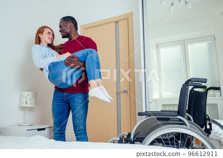 Young bearded handicapped redhaired ginger wife and her african american husband in bedroom. man holds her at hands and helping to reach a bed Young bearded handicapped redhaired ginger wife and her african american husband in bedroom. man holds her at hands and helping to reach a bed 96612911