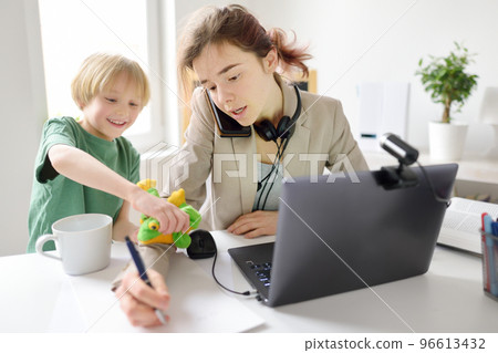 Young woman working online using laptop at home. Home office and parenthood at same time. Exhausted parent with hyperactive child. Mischievous kid of mother freelancer. 96613432