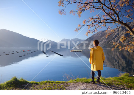 Young woman in yellow coat standing on the coast of Adriatic sea and admiring of stunning winter view of Boka Kotor Bay seascape. Tourism and travel in Montenegro. 96613465