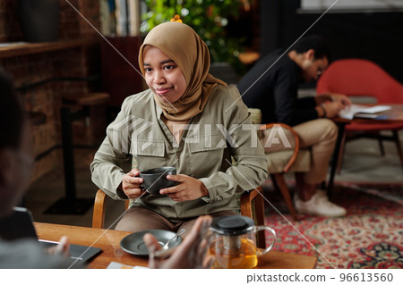 Young Muslim female employee with cup of herbal tea looking at colleague Young Muslim female employee with cup of herbal tea looking at colleague 96613560