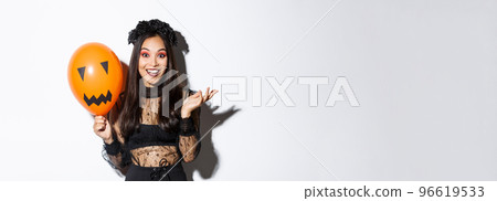 Image of beautiful asian woman celebrating halloween, wearing witch costume and gothic makeup, talking with orange balloon 96619533