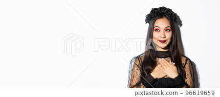 Close-up of nostalgic smiling asian woman looking at upper left corner dreamy, holding hands on heart, wearing gothic lace dress for halloween party 96619659