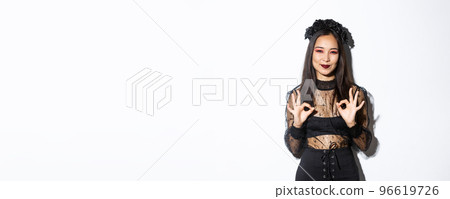 Image of smiling asian cute witch wearing black gothic dress and wreath showing okay gestures in approval, praise something, standing in halloween costume against white background Image of smiling asian cute witch wearing black gothic dress and wreath showing okay gestures in approval, praise something, standing in halloween costume against white background 96619726