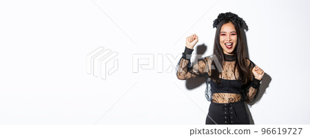 Image of young asian woman having fun on halloween party, raising hand up as rejoicing or triumphing, wearing witch gothic dress, standing over white background Image of young asian woman having fun on halloween party, raising hand up as rejoicing or triumphing, wearing witch gothic dress, standing over white background 96619727