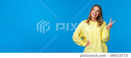 Staying bright sie. Carefree happy young silly blond girl in yellow hoodie sending positive vibes, show kawaii peace sign and smiling with white teeth, standing blue background Staying bright sie. Carefree happy young silly blond girl in yellow hoodie sending positive vibes, show kawaii peace sign and smiling with white teeth, standing blue background 96619733