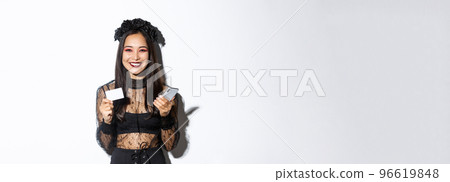 Beautiful stylish asian woman in halloween costume holding mobile phone with credit card and smiling, standing over white background Beautiful stylish asian woman in halloween costume holding mobile phone with credit card and smiling, standing over white background 96619848
