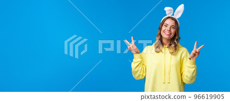 Holidays, traditions and celebration concept. Kawaii young blond girl in rabbit ears showing peace gesture and smiling, having fun, enjoying party, standing blue background 96619905