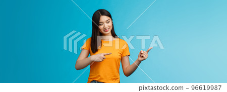 Tender lovely asian girl with dark haircut look curiously pointing left, interested in new product in store, stand blue background amused, entertained, observe exhibition, wear yellow t-shirt 96619987