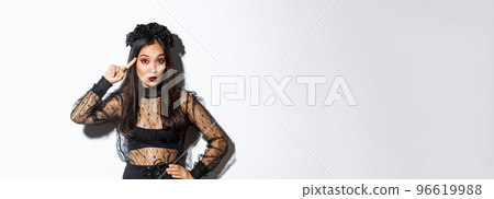 Image of annoyed young woman in halloween gothic dress scolding someone stupid, rolling finger on temple and looking at crazy person, standing over white background 96619988