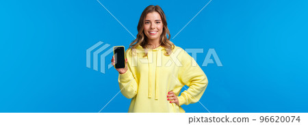 Waist-up portrait good-looking blond girl in yellow hoodie promote smartphone application, show mobile phone display and smiling camera as recommend subscribe or download app 96620074