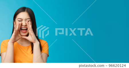 Carefree asian enthusiastic girl shouting long distance, calling friend, breaking free emotions, yelling out loud, hold hands near opene mouth rupor, searching someone in crowd, blue background 96620124