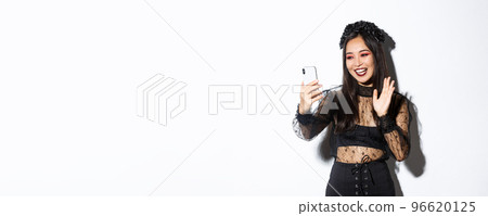 Portrait of beautiful and stylish asian woman in gothic lace dress saying hi, waving hand at smartphone camera during video call, standing over white background 96620125
