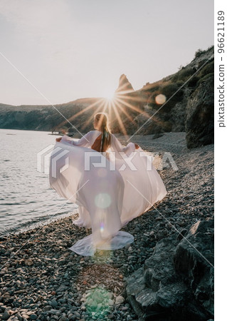 Mysterious young woman with braids in long white dress alone on the beach. Sunset over the sea with rocky volcanic cliff. Abstract nature summer ocean sea background. 96621189