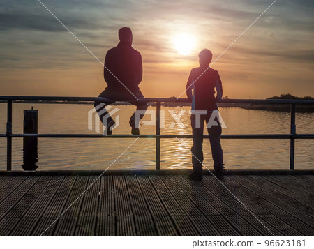 Couple man and woman looks at sunset over the sea. Man sits on a railing Couple man and woman looks at sunset over the sea. Man sits on a railing 96623181