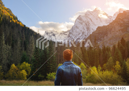 Back view of a man in a knitted hat and denim jacket against the backdrop of a green dense forest and snow-capped high mountains Back view of a man in a knitted hat and denim jacket against the backdrop of a green dense forest and snow-capped high mountains 96624495