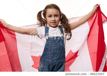 Happy little girl with two ponytails, wearing blue denim overalls, carries Canadian flag. Concept of Independence Day of Canada, the first July. Immigration. Emigration. Travel and tourism concept Happy little girl with two ponytails, wearing blue denim overalls, carries Canadian flag. Concept of Independence Day of Canada, the first July. Immigration. Emigration. Travel and tourism concept 96625947