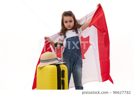 Cute little girl in white t-shirt and blue denim overalls, carrying Canada flag, smiles looking at camera while posing with a yellow suitcase over white background with free space for advertising text Cute little girl in white t-shirt and blue denim overalls, carrying Canada flag, smiles looking at camera while posing with a yellow suitcase over white background with free space for advertising text 96627782