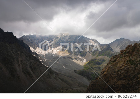 Dramatic view of forested high rocky slopes and snowy mountains in gray cloudy sky in changeable weather, mountain landscape without people Dramatic view of forested high rocky slopes and snowy mountains in gray cloudy sky in changeable weather, mountain landscape without people 96628274