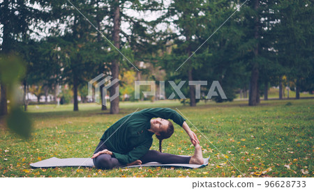 Smiling young woman is exercising in park doing yoga sitting on mat on green and yellow grass enjoying fresh air and healthy activity. Youth, active lifestyle and recreation concept. Smiling young woman is exercising in park doing yoga sitting on mat on green and yellow grass enjoying fresh air and healthy activity. Youth, active lifestyle and recreation concept. 96628733