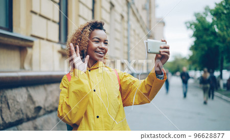 Cute African American teenager is talking to friends onlin using smartphone and looking at screen standing outdoors in the street of modern city and wearing earphones. Cute African American teenager is talking to friends onlin using smartphone and looking at screen standing outdoors in the street of modern city and wearing earphones. 96628877