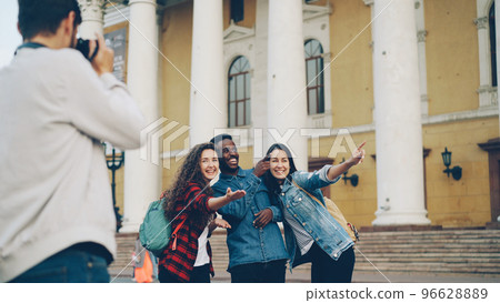 Joyful tourists men and women are posing for camera with beautiful building in background having fun laughing and jumping while bearded guy is holding camera and shooting. 96628889