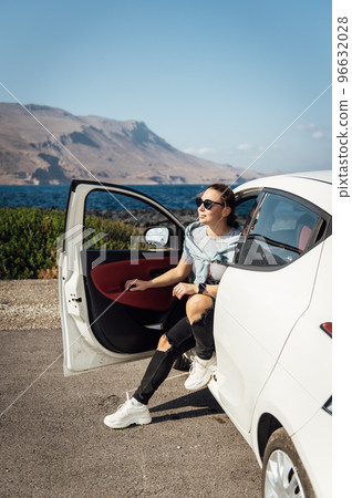 A beautiful brunette in sunglasses sits by a white car against the backdrop of the Mediterranean Sea and mountains A beautiful brunette in sunglasses sits by a white car against the backdrop of the Mediterranean Sea and mountains 96632028