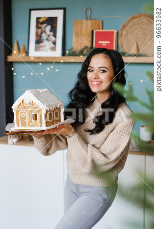 A smiling, happy, attractive young woman holds a Christmas gingerbread house, which she baked herself, in the Scandinavian-style kitchen 96633982