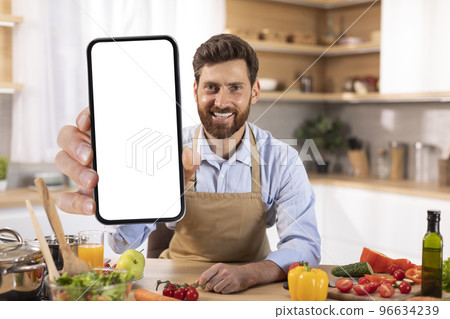 Happy adult caucasian male with beard in apron shows smartphone with blank screen in modern kitchen interior 96634239