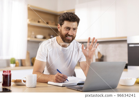 Smiling mature caucasian man with beard in white t-shirt, waving hand at laptop webcam, make notes 96634241