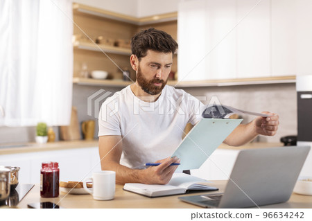 Serious adult caucasian male with beard works with documents and laptop in minimalist kitchen interior 96634242