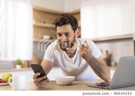 Smiling mature caucasian male with beard in white t-shirt eats porridge, read news on smartphone, watches video Smiling mature caucasian male with beard in white t-shirt eats porridge, read news on smartphone, watches video 96634243