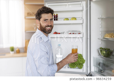 Happy handsome adult caucasian man with beard in shirt opens refrigerator door, takes glass of juice Happy handsome adult caucasian man with beard in shirt opens refrigerator door, takes glass of juice 96634259