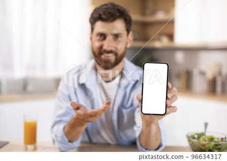 Portrait of glad adult caucasian man with beard shows phone with blank screen in kitchen interior Portrait of glad adult caucasian man with beard shows phone with blank screen in kitchen interior 96634317