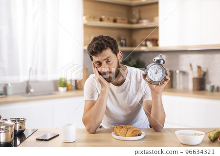 Sad tired middle aged caucasian male with beard in white t-shirt shows alarm clock and sleeps in modern kitchen 96634321
