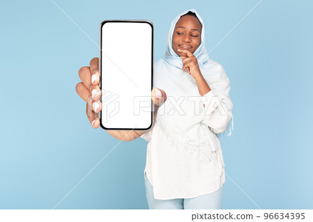 Happy pensive black muslim woman thinking, showing huge smartphone with blank screen, blue background, mockup 96634395
