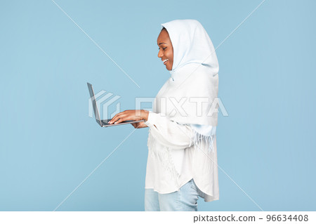 African american muslim woman typing on computer keyboard and smiling over blue studio background, side view, copy space African american muslim woman typing on computer keyboard and smiling over blue studio background, side view, copy space 96634408