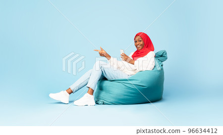Happy black muslim woman sitting at bean bag with smartphone and pointing at copy space, showing nice offer 96634412