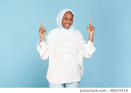 Excited african american muslim woman pointing up above her head with two hands, demonstrating free place for your ad 96634419