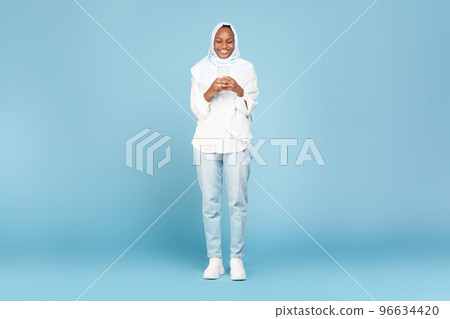 Full length shot of happy black muslim woman using smartphone, checking mobile application, blue background 96634420