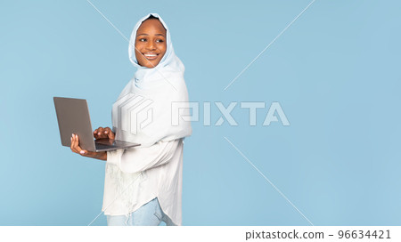Smiling black woman in hijab standing with laptop computer and looking at free space over blue studio background 96634421