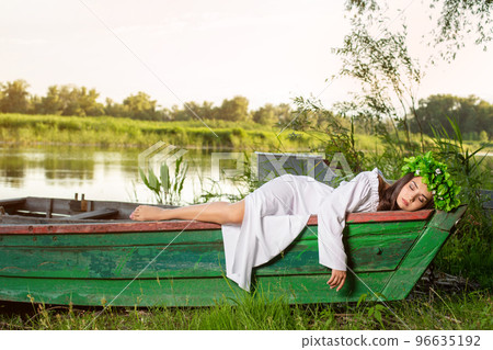 The nymph with long dark hair in a white vintage dress sitting in a boat in the middle of the river. 96635192