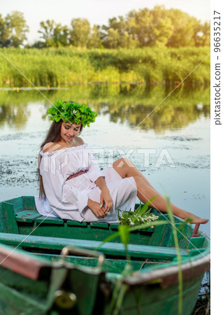 The nymph with long dark hair in a white vintage dress sitting in a boat in the middle of the river. 96635217