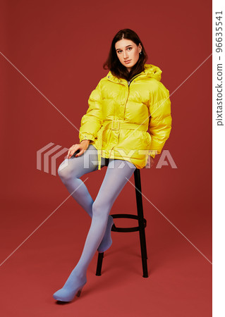 Beautiful young fashion woman in yellow oversized down jacket posing in studio on red background, using bar stool 96635541