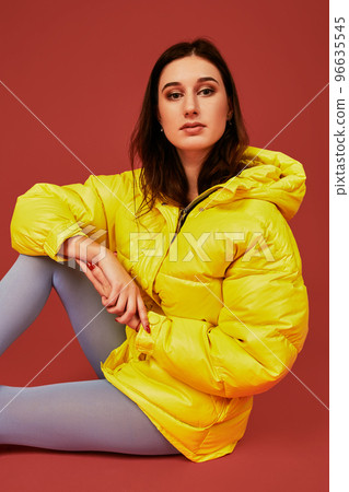 Studio portrait of young brunette woman sitting on the floor in yellow down jacket and grey blue panty hoses or stockings Studio portrait of young brunette woman sitting on the floor in yellow down jacket and grey blue panty hoses or stockings 96635545