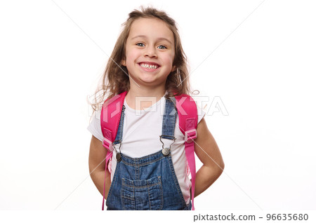Isolated portrait on white background of a Caucasian beautiful little school child, a baby girl wearing blue denim overalls, with pink backpack, cutely smiling cheerful toothy smile looking at camera Isolated portrait on white background of a Caucasian beautiful little school child, a baby girl wearing blue denim overalls, with pink backpack, cutely smiling cheerful toothy smile looking at camera 96635680
