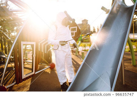 Man wearing protective suit disinfecting the playground in the sun with spray. Covid -19. Cleaning. Man wearing protective suit disinfecting the playground in the sun with spray. Covid -19. Cleaning. 96635747