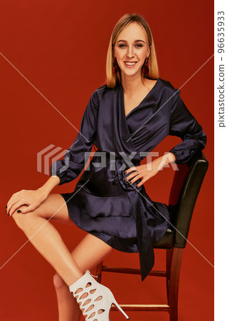 Beautiful young blonde woman in elegant black cocktail dress is posing for camera leaning on a chair 96635933